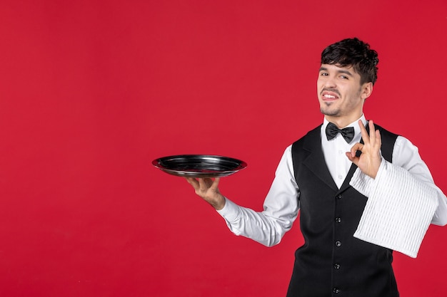 Vista superior de um jovem sorridente garçom de uniforme com gravata borboleta segurando a bandeja e uma toalha, fazendo um gesto perfeito