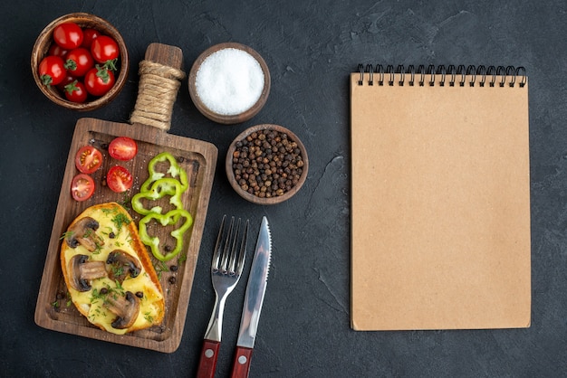 Vista superior de um lanche saboroso caseiro com cogumelos e legumes picados em uma tábua de madeira, talheres, conjunto de caderno de especiarias em fundo preto