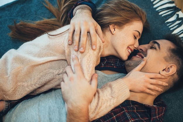 Vista superior de um lindo casal jovem rindo enquanto se abraça cara a cara, inclinando-se na cama enquanto a garota está tocando o rosto do namorado.