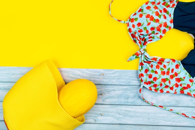Vista superior de um maiô e um melão em uma bolsa, sobre um fundo azul de madeira e amarelo. Conceito de férias de verão.