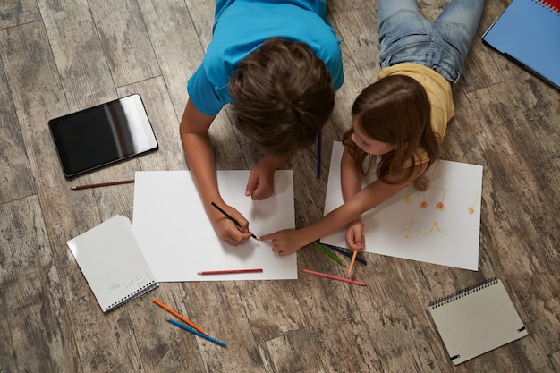 Vista superior de um menino caucasiano e uma menina deitada no chão de madeira em casa e desenhando em um branco