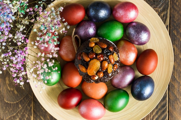 Vista superior de um prato com um bolo de Páscoa e ovos brilhantes pintados. flores da primavera. fundo de madeira.