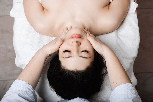 Vista superior de uma bela jovem mulher apoiada em uma cama de spa com os olhos fechados, relaxando enquanto faz uma massagem facial por um cosmetologista em um centro de spa.