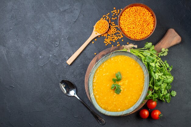 Vista superior de uma deliciosa sopa em uma tigela na placa de madeira um molho de tomates verdes e lentilhas vermelhas no lado esquerdo em fundo preto