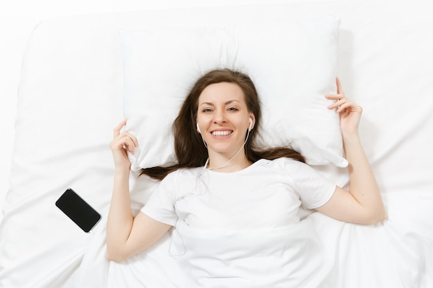 Foto vista superior de uma jovem feliz deitada na cama com um lençol branco, travesseiro, cobertor, ouça música com fones de ouvido no celular