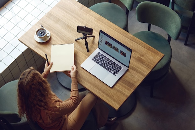 Vista superior de uma jovem mulher casual escrevendo notas enquanto está sentada à mesa de madeira com o laptop