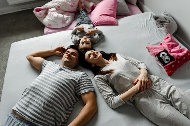 Vista superior de uma linda garotinha e seus lindos pais jovens olhando para a câmera e sorrindo, deitada no chão em casa, Vvew de cima de uma família descansando deitada no chão e olhando para a câmera