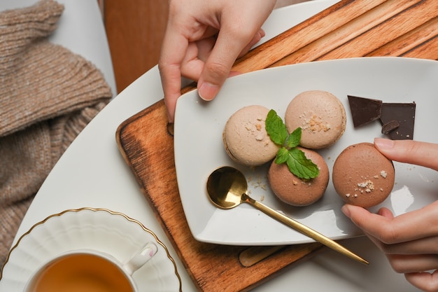 Vista superior de uma mão feminina colhendo macarons franceses em um prato na mesa de centro com uma xícara de chá