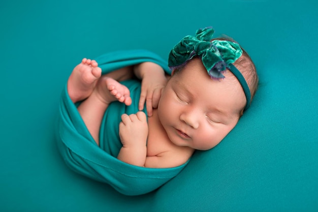 Vista superior de uma menina recém-nascida dormindo em um envoltório turquesa com uma faixa turquesa e uma flor na cabeça em um fundo turquesa Belo retrato de uma menina 7 dias por semana