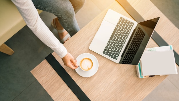 Vista superior de uma mulher asiática usando laptop na mesa de madeira, sentado e segurando uma xícara de café com leite quente enquanto está sentado no sofá. mulher de negócios, trabalhando no laptop em casa.