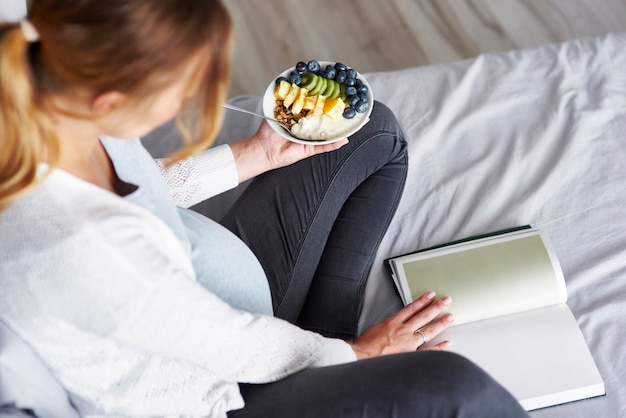 Vista superior de uma mulher grávida comendo e lendo um livro