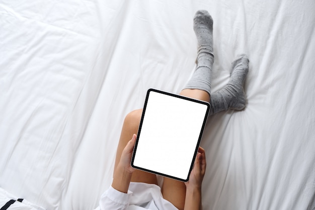 Vista superior de uma mulher segurando preto tablet pc com tela branca de mesa em branco, enquanto está sentado em uma cama branca aconchegante em casa