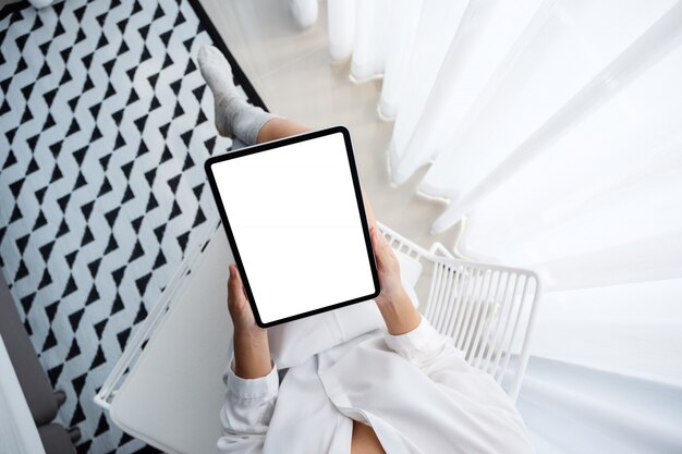 Vista superior de uma mulher segurando preto tablet pc com tela de mesa branca em branco enquanto está sentado no quarto com sensação de relaxamento de manhã