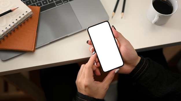 Vista superior de uma mulher usando maquete de tela branca de smartphone moderno sobre o espaço de trabalho moderno