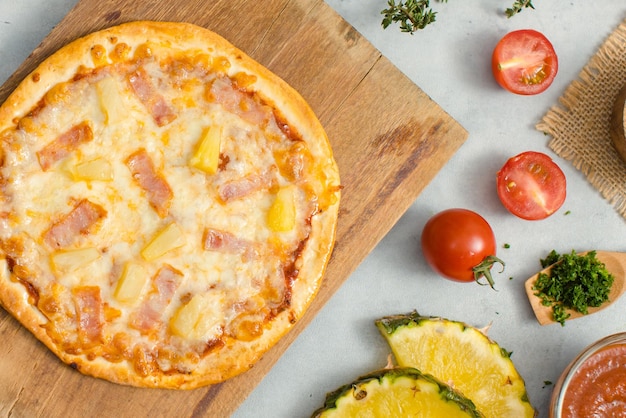 Vista superior de la deliciosa y sabrosa pizza de piña y jamón crujiente de corteza italiana tradicional hawaiana colocada en una tabla de cortar de madera con ingredientes tomates en rodajas ketchup y sal