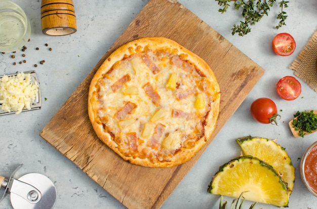 Vista superior de la deliciosa y sabrosa pizza de piña y jamón crujiente de corteza italiana tradicional hawaiana colocada en una tabla de cortar de madera con ingredientes tomates en rodajas ketchup y sal