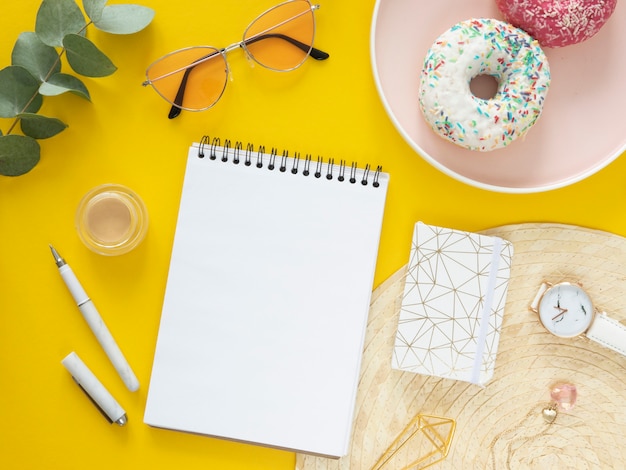 Vista superior Desayuno femenino en el trabajo. Maqueta de cuaderno espiral plano, papelería y donas sobre un fondo amarillo