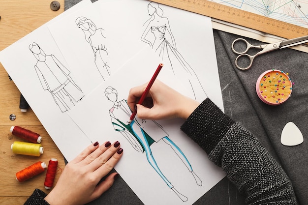 Vista superior del diseñador de moda en el trabajo. Manos femeninas dibujando ropa boceto en su espacio de trabajo creativo, vista superior