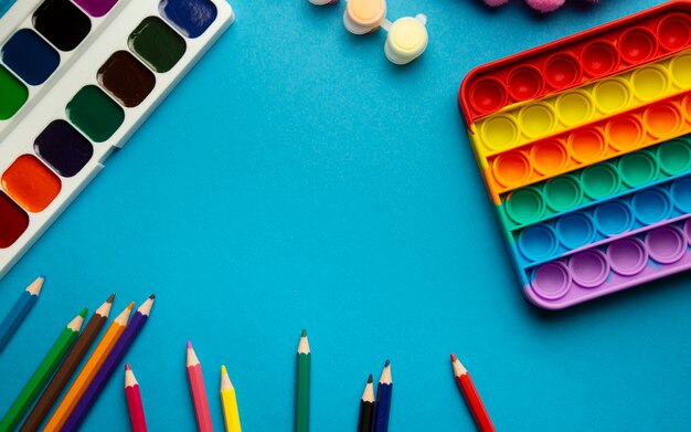 Vista superior de diseño de artículos escolares para niños, pop it, lápices de colores de acuarelas de juguete sobre un fondo azul con lugar para texto, útiles escolares de colores