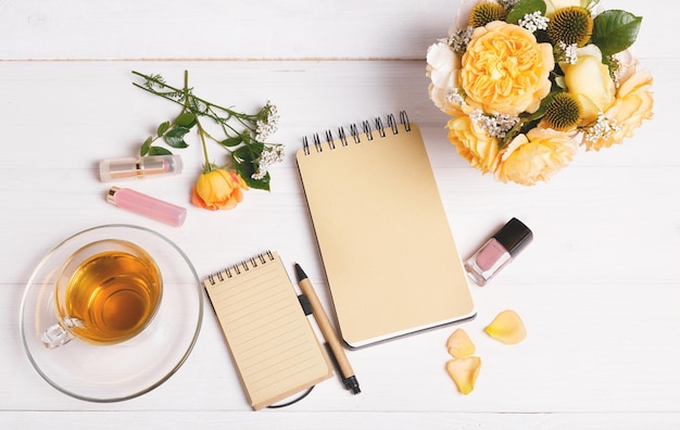 Vista superior do caderno aberto vazio com caneta flores inglês rosa cosméticos e xícara de chá no topo da mesa