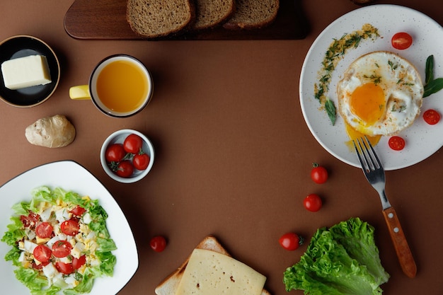 Vista superior do café da manhã com ovo frito com tomate e endro e salada de legumes com tomate ovo alface com garfo suco de laranja fatias de pão manteiga alface no fundo marrom