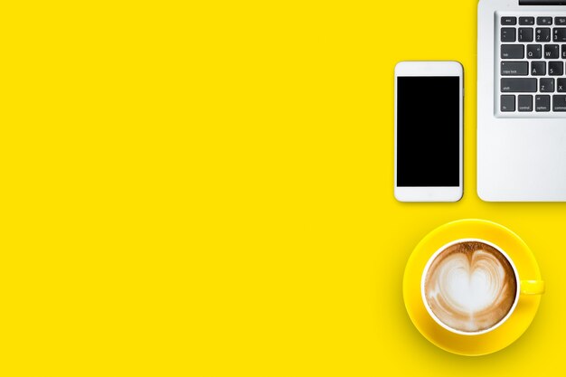 Vista superior do café latte ou cappuccino em copo amarelo com smartphone e laptop com espaço de cópia na mesa amarela