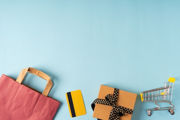 Vista superior do carrinho de compras de brinquedos, caixas, cartão de crédito, saco de papel preto e um presente sobre fundo azul.