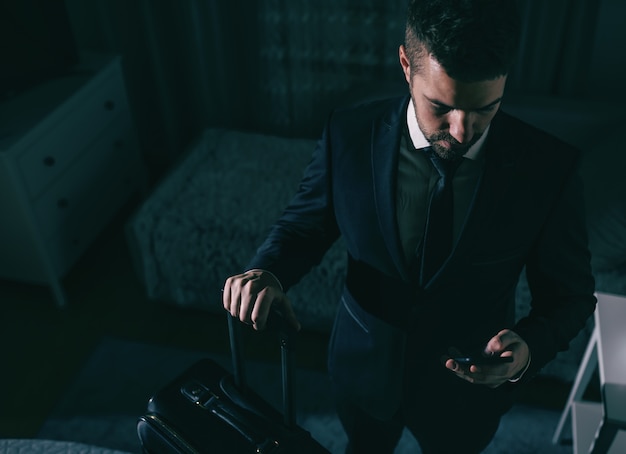 Vista superior do empresário em terno de pé no quarto, segurando a mala e usando o telefone inteligente. conceito de viagem de negócios.