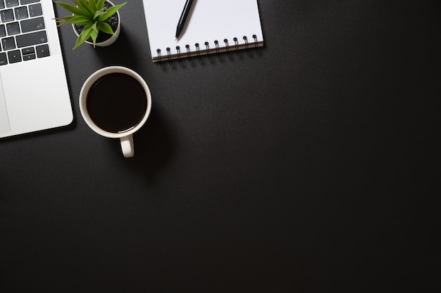 Vista superior do escritório de mesa com computador portátil, bloco de notas em branco, caneta, café e planta em preto