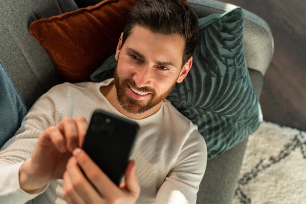 Vista superior do homem caucasiano digitando no teclado do smartphone enquanto estava deitado no sofá e trabalhando em casa ou conversando com amigos