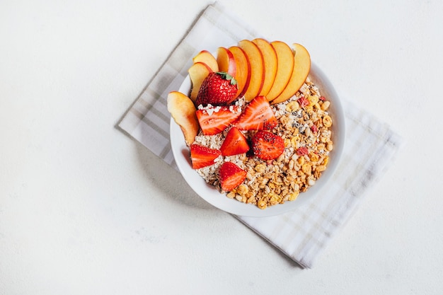 Vista superior do mingau de aveia no café da manhã com flocos de granola, frutas, morangos e pêssegos em um prato branco sobre um fundo branco