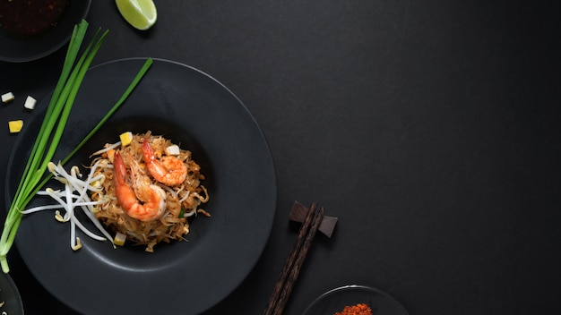 Vista superior do pad thai, agite a mosca de macarrão tailandês com camarão, ovo e tempero em chapa de cerâmica preta na mesa preta