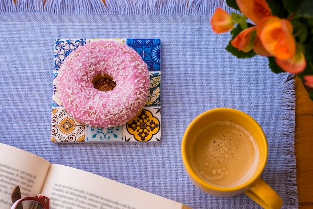 Vista superior de donut helado rosa dulce en la mesa de madera taza de café y flores de naranja pausa o desayuno con un libro