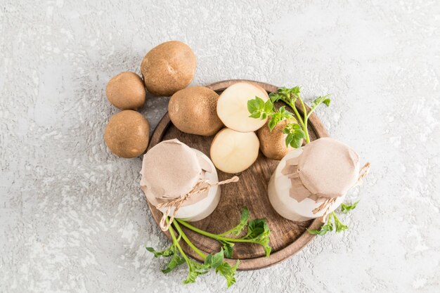 Vista superior de dos botellas de vidrio con leche de patata vegetal y tubérculos de patata picada sobre una bandeja de madera fondo de hormigón gris