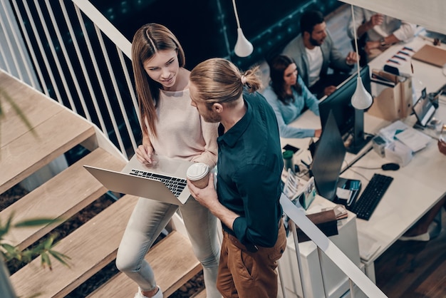 Vista superior de dos jóvenes colegas modernos en ropa casual elegante que se comunican y usan la computadora portátil mientras están de pie en las escaleras