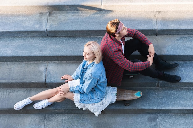 La vista superior de la elegante pareja de adolescentes con un longboard está sentada en una escalera de hormigón