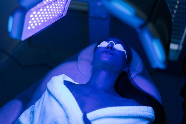 Vista superior em close-up de uma cliente feminina que está a receber um tratamento de terapia fotodinâmica facial com luz led azul em