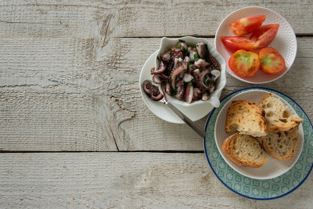 Vista superior de ensalada de pulpo con rodajas de baguette tostado y tomates sobre fondo de madera rústica. Aperitivo típico mediterráneo