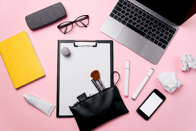 Vista superior del escritorio de oficina rosa de la mujer con laptop, teléfono con pantalla blanca, anteojos, lápiz labial, crema, bolsa de cosméticos y fondo de bolas de papel arrugado.