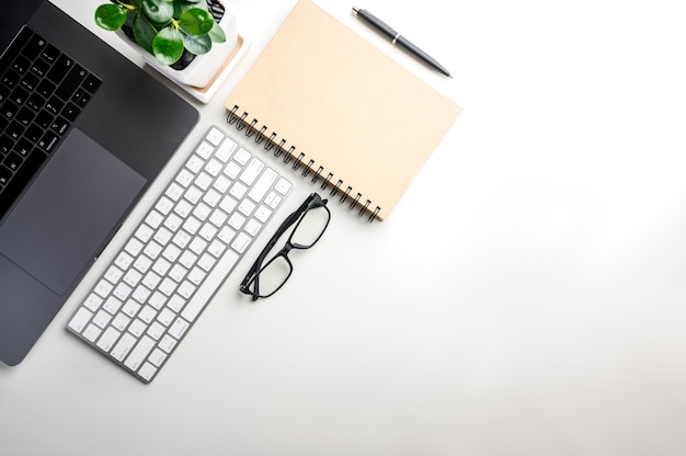 Foto vista superior con espacio de copia de mesa blanca con teléfono inteligente, computadora portátil, teclado, cuaderno, bolígrafo, árbol pequeño y gafas.