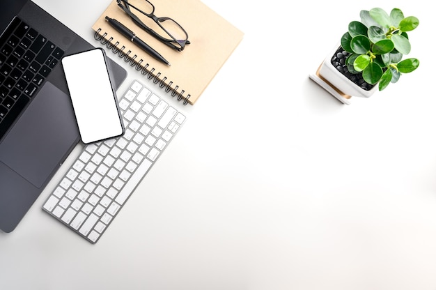 Foto vista superior con espacio de copia de mesa blanca con teléfono inteligente, computadora portátil, teclado, cuaderno, bolígrafo, árbol pequeño y gafas.