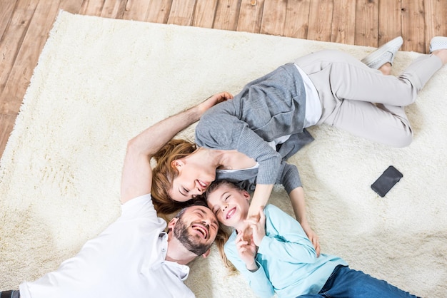 Vista superior de la familia de tres relajándose en casa acostados en la alfombra y riendo