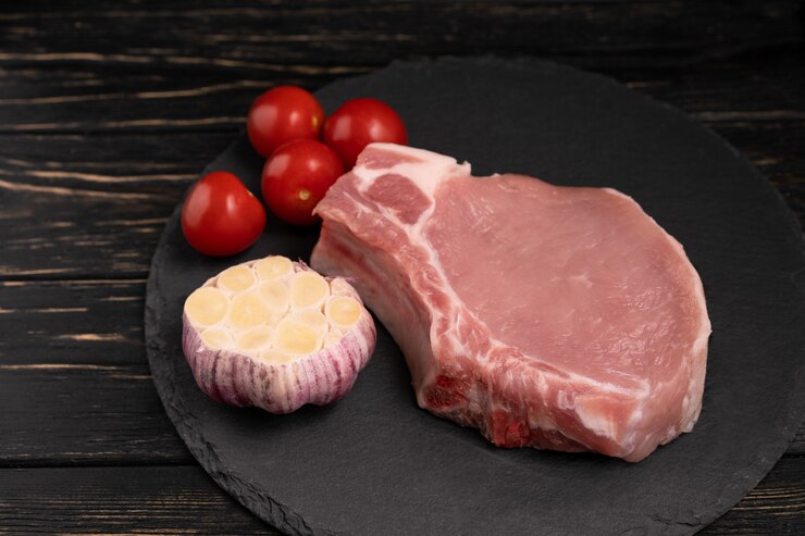 Vista superior de filetes de chuleta de cerdo cruda de una pieza con tomates cherry y ajo en una tabla de cortar de piedra negra.