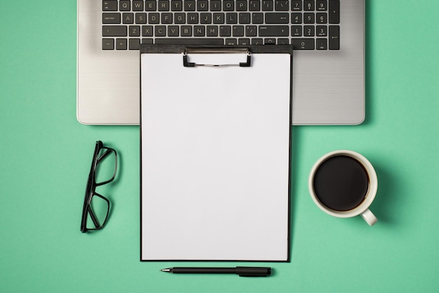 Vista superior de la foto de la carpeta del portapapeles negro con una hoja de papel blanco en el teclado del portátil, gafas de lápiz y una taza de café en un fondo turquesa aislado con espacio de copia