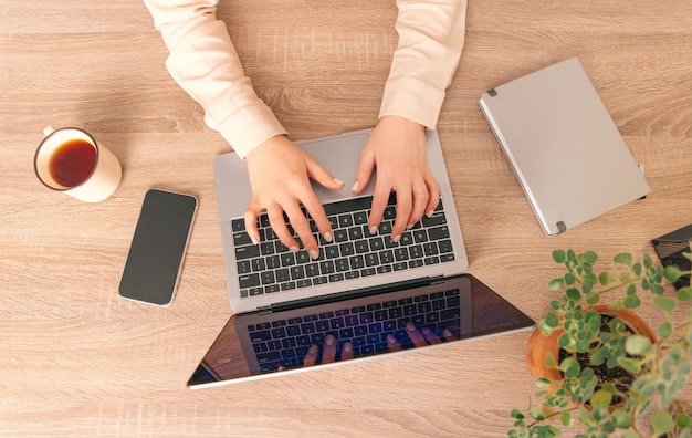 Vista superior de la foto de un escritorio de trabajo de una mujer escribiendo en la computadora portátil La taza del teléfono de la planta y el diario también están en el escritorio