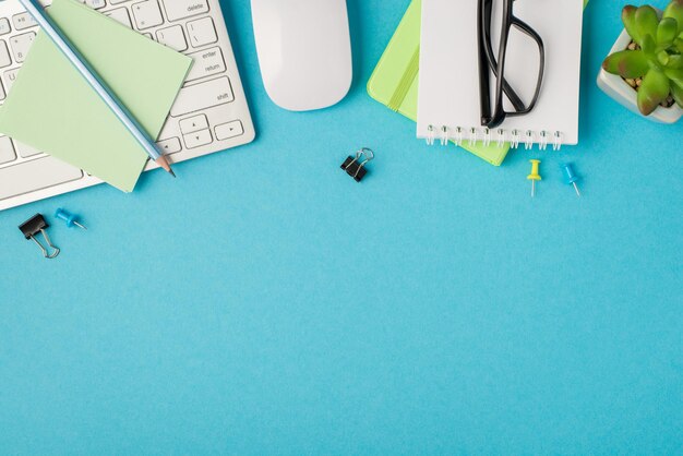 Vista superior de la foto del lugar de trabajo con la pegatina de la cancillería del teclado, clips de lápiz, gafas de ratón en cuadernos y planta sobre fondo azul aislado con espacio de copia