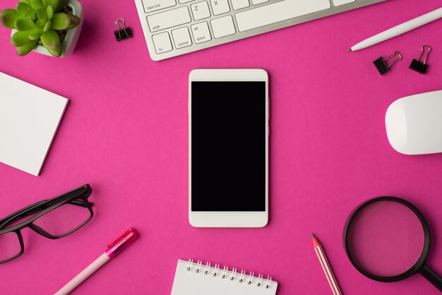 Vista superior de la foto del lugar de trabajo con el teléfono inteligente en el medio teclado mouse cancillería lupa planta gafas y cuadernos sobre fondo rosa aislado con copyspace