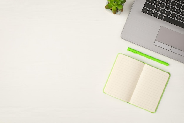 Vista superior de la foto de la maceta de la computadora portátil gris, el planificador abierto verde claro y la pluma sobre el fondo de la mesa de madera blanca aislada con espacio en blanco