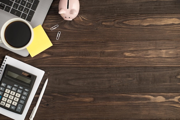 Vista superior de la foto de la taza de bebida etiqueta amarilla papel de nota en la computadora portátil alcancía clips pluma y calculadora en el planificador en el fondo aislado de la mesa de madera oscura con espacio vacío