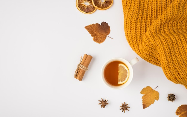 Vista superior de la foto de la taza de té de suéter de punto amarillo con hojas de morera de otoño amarillo limón, palitos de canela de anís y rodajas de limón seco sobre fondo blanco aislado con espacio de copia